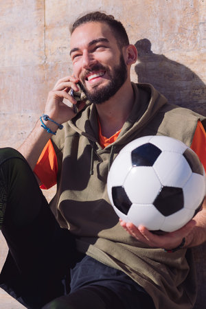 handsome sportsman laughing and talking by phone while resting with his football ball, concept of...