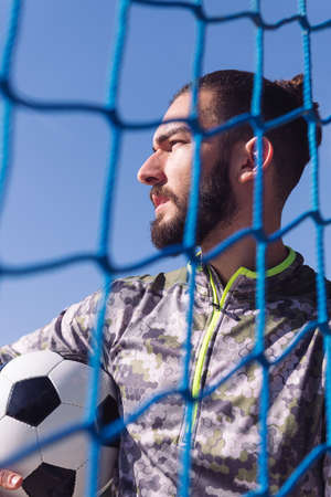 vertical portrait of an attractive sportsman through the goal net with the football ball under his arm, concept of healthy lifestyle and urban sport in the cityの写真素材