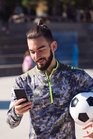 fashionable sportsman with a soccer ball smiles consulting his mobile phone on the football court,...
