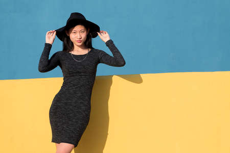 fashionable asian young woman posing in a black tight dress and hat on a colorful yellow and blue...