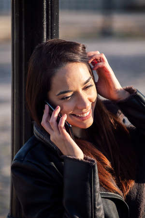 vertical photo of a smiling young woman talking on the phone leaning against a lamppost, concept of...