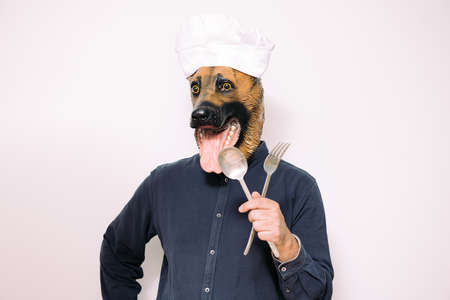 person with a dog mask and chef's hat showing eating utensils on white background, concept of care and balanced feeding for petsの写真素材