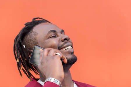 portrait of a stylish black businessman laughing and talking on phone in front of red background,...