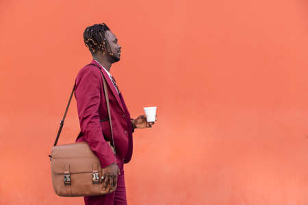 elegant african businessman in a suit with briefcase holding a coffee in his hand in front of a red...