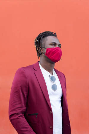 vertical photo of a stylish black man with protective mask to match his suit on a red background, concept of elegance and fashionable lifestyle, copy space for textの写真素材
