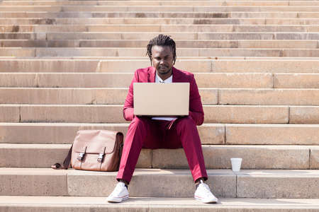 elegant black businessman works with his laptop sitting on a staircase in the city, technology and...