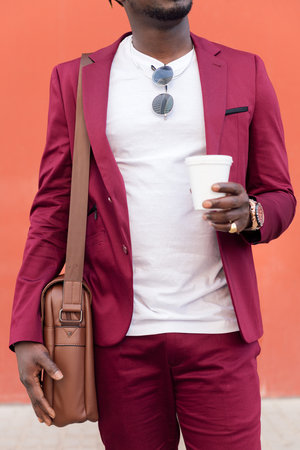 vertical photo of an unrecognizable elegant black businessman dressed in suit with briefcase holds coffee in his hand in front of red background, copy space for textの写真素材