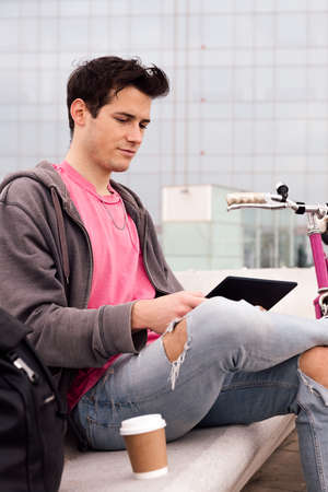vertical photo of a young student sitting outdoors using tablet next to his bicycle, concept of technology and urban lifestyle, copy space for textの写真素材