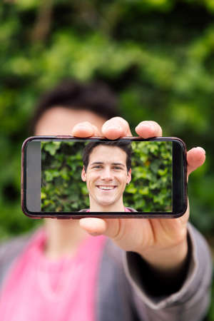 vertical photo of a happy young man covering his face with a smart phone with a photo of himself,...