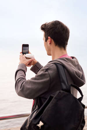 rear view of a young student with backpack and coffee taking a photo with a mobile phone, concept of technology and millennial lifestyle, copy space for text, vertical photoの写真素材