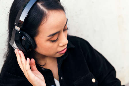 portrait of a young asian woman with headphones enjoying listening to music outdoors leaning against a concrete wallの写真素材