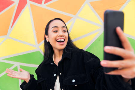 happy young asian woman having fun by making a video call with her mobile phone next to a colorful wallの写真素材