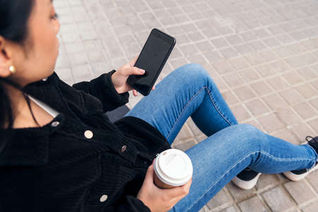 unrecognizable asian girl typing on a phone sitting outdoors on a concrete floorの写真素材