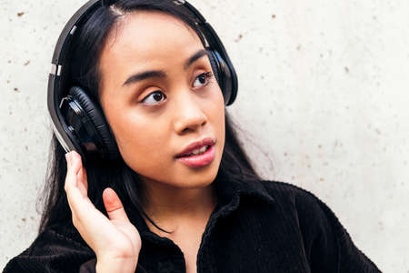 young asian woman enjoys listening to music with headphones outdoors leaning against a concrete wallの写真素材