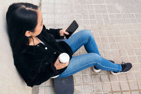 unrecognizable asian girl typing on a phone sitting outdoors on her skateboard leaning against a concrete wallの写真素材