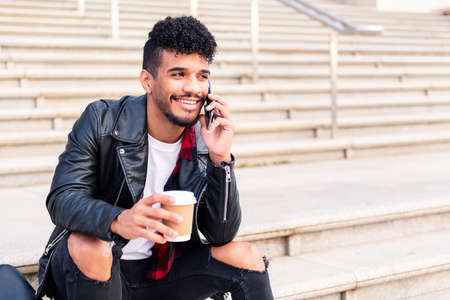 happy young latin student sitting on a staircase smiles talking on the phone with a coffee in his...