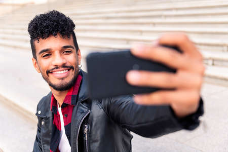 happy young latin man smiles as he takes a selfie with his smart phone, concept of technology and urban lifestyleの写真素材