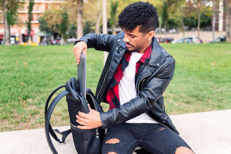 young latin man sitting on a city bench takes a laptop out of his backpack, concept of technology and urban lifestyleの写真素材