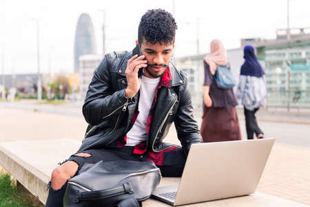 stylish latin man typing on his computer while talking on the phone sitting in the city as two arab women walk past him, concept of technology and urban lifestyleの写真素材