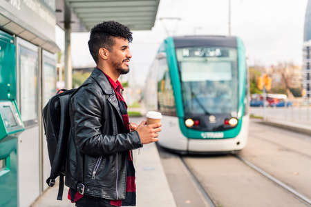 stylish young latin man with a take away coffee in his hand waits at the public transport stop for the arrival of the streetcar, concept of urban lifestyleの写真素材