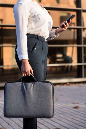unrecognizable businesswoman with briefcase consulting her phone in front of office building, concept of successful entrepreneur and urban lifestyleの写真素材