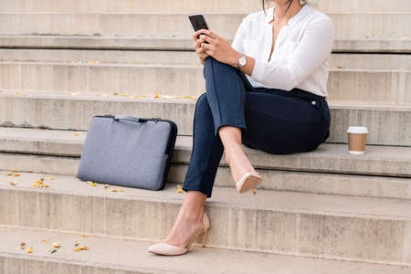 unrecognizable businesswoman consulting her phone sitting with legs crossed on concrete stairs, concept of digital entrepreneur and urban lifestyleの写真素材