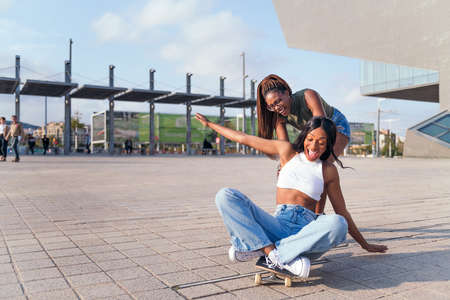 two young black female friends laugh in amusement playing with a skateboard, concept of youth and friendship, copy space for textの写真素材