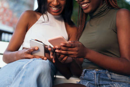 detail of the hands of two unrecognizable smiling black women looking at their mobile phones, concept of youth and communicationの写真素材