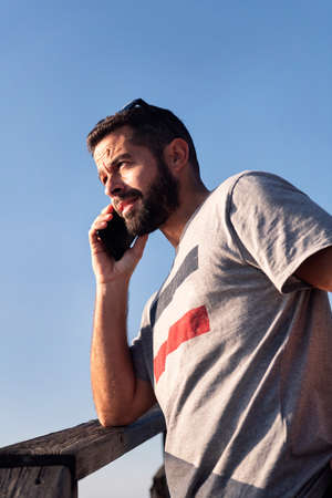 vertical portrait of a handsome young man talking on his smart phone leaning on a wooden railing...