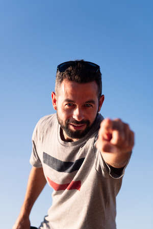 vertical photo of a bearded man pointing finger at camera with blue sky in background, copy space...