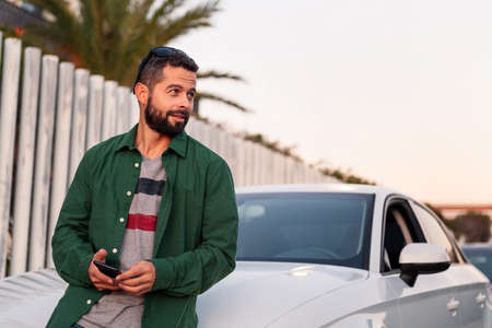 attractive young bearded man sitting on the hood of his car with mobile phone in hand, leisure and...
