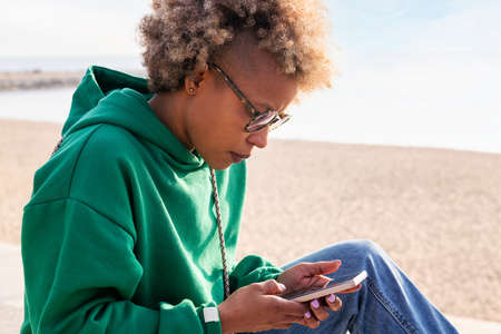 young latin woman in a hoodie using her mobile phone next to the beach, concept of lifestyle and...