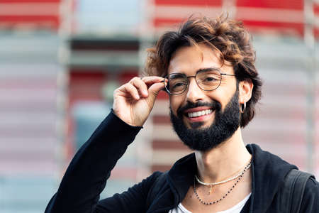 portrait of a smiling young man with beard putting on his glasses, concept of youth and happiness,...