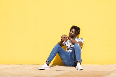african man sitting at floor against yellow wallの写真素材