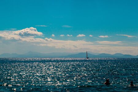 seascape formed by 2 bathers and a sailboat, in the background a mountainous island is intulledの写真素材