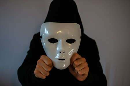 Man with black hood holding an anonymous white mask on dark backgroundの写真素材