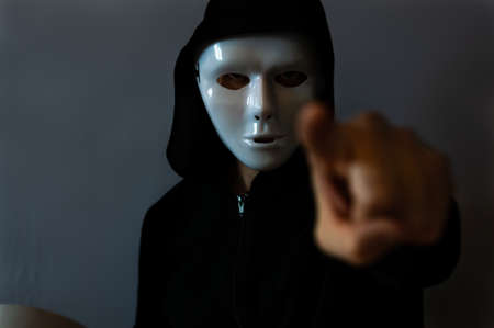 Man with a black hooded jacket wearing a halloween white mask and with a pointing finger, on dark backgroundの写真素材