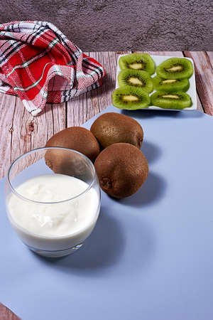 Glass with yoghurt and broken kiwis on a plate. Wooden floor and wooden board, whole kiwis, white and red kitchen towel, blue mat.の写真素材