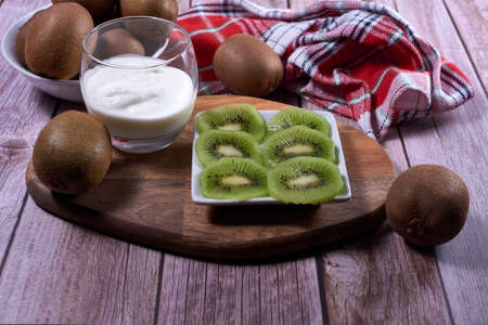 Yoghurt glass and plate of cut kiwis and several whole ones on wood. Wooden board, red and white kitchen towel, squares, white bowl, front view.の写真素材