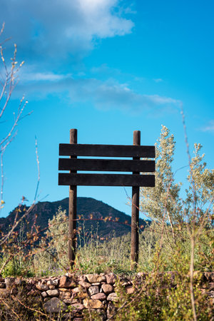 A vertical medium shot features a rustic, dark wooden signpost with three horizontal planks, positioned centrally against a bright blue sky with scattered white clouds. The sign is supported by two vertical posts. The foreground displays dry grass, small green bushes, and a traditional stone wall. A large mountain is visible in the background, implying a rural or natural park setting. This image provides ample copy space for text.の写真素材