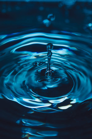 328.Droplet impacts blue-tinted water forming vertical splash and concentric ripples. Lighting...