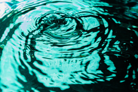 Close-up of water surface showing vortex and ripple rings. Teal lighting creates reflections and shadows. Image captures fluid motion, wave behavior, and surface tension in detail.の写真素材
