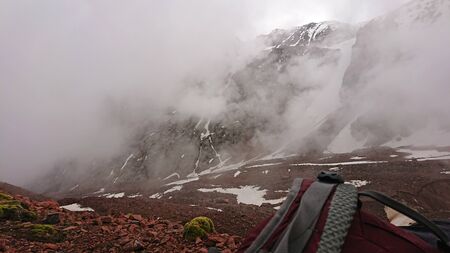 View of the strap backpack in the mountains. Clouds drift past. Snow is visible on the mountain slopes. Green moss grows on the stones. Hiking in the mountains.の写真素材