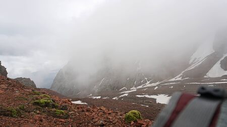 View of the strap backpack in the mountains. Clouds drift past. Snow is visible on the mountain slopes. Green moss grows on the stones. Hiking in the mountains.の写真素材