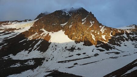 The sun falls on the summit of the peak. Clouds are visible. On rocky terrain lies snow. The shadow of the mountains.の写真素材