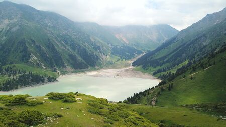 Big Almaty lake located in the mountains of Kazakhstan. It offers views of green grass, flowers, lake, rocks, large mountains and the sky in the clouds. Mountain lake with blue water.の写真素材