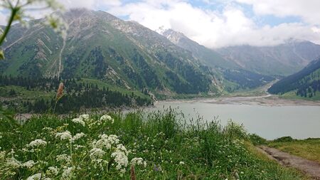 Big Almaty lake located in the mountains of Kazakhstan. It offers views of green grass, flowers, lake, rocks, large mountains and the sky in the clouds. Mountain lake with blue water.の写真素材