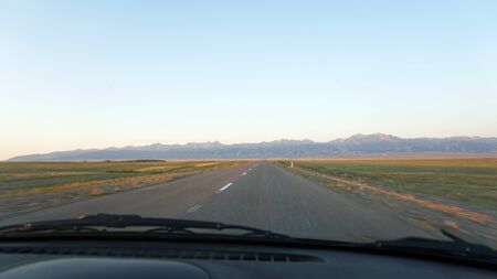 View of the road from the car. Straight line. Hills on the horizon. The car rides on the steppe, on the sides of the road grass grows. In the distance you can see the blue sky.の写真素材
