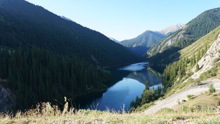 Kolsay lake located in the mountains of Kazakhstan. Turquoise water as a mirror. The water reflects the mountains, green hills, tall spruce, grass, sky and clouds. Beautiful landscape of mountain lake.の写真素材