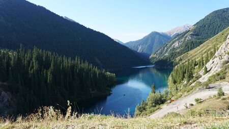 Kolsay lake located in the mountains of Kazakhstan. Turquoise water as a mirror. The water reflects the mountains, green hills, tall spruce, grass, sky and clouds. Beautiful landscape of mountain lake.の写真素材